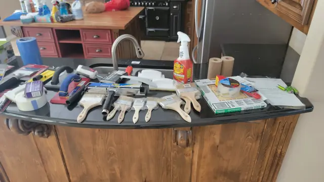 New paintbrushes, ceiling scraper, sanding sponges and prep supplies laid out on a granite counter