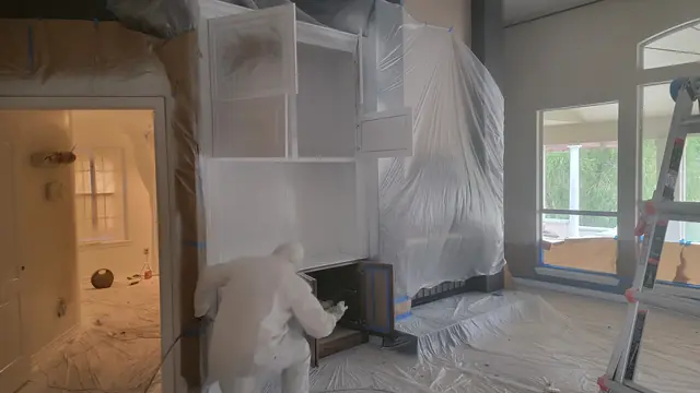 A worker in protective coveralls spray-refinishing built-in cabinets white