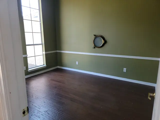 An empty room before renovation with olive-green textured walls, chair rail and dark hardwood floors